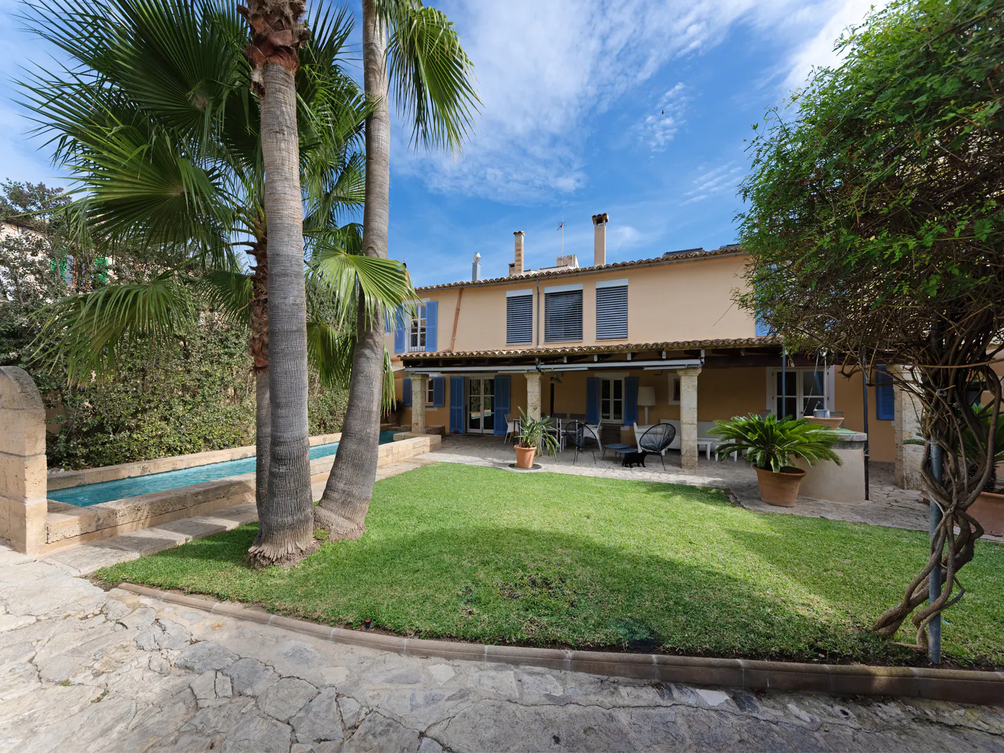 Backyard view of a two-story beige house with blue shutters, a green lawn, palm trees, and a pool.