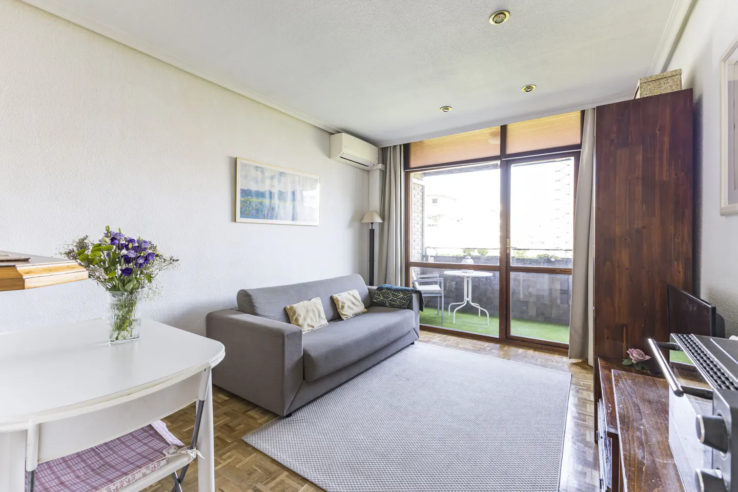 Bright living room with a gray sofa, white table with flowers, and a balcony with a table and chairs.