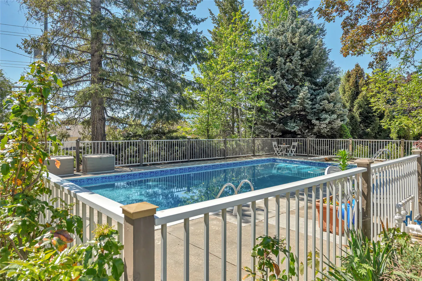 Outdoor pool with blue water, surrounded by a white fence and lush green trees on a sunny day.