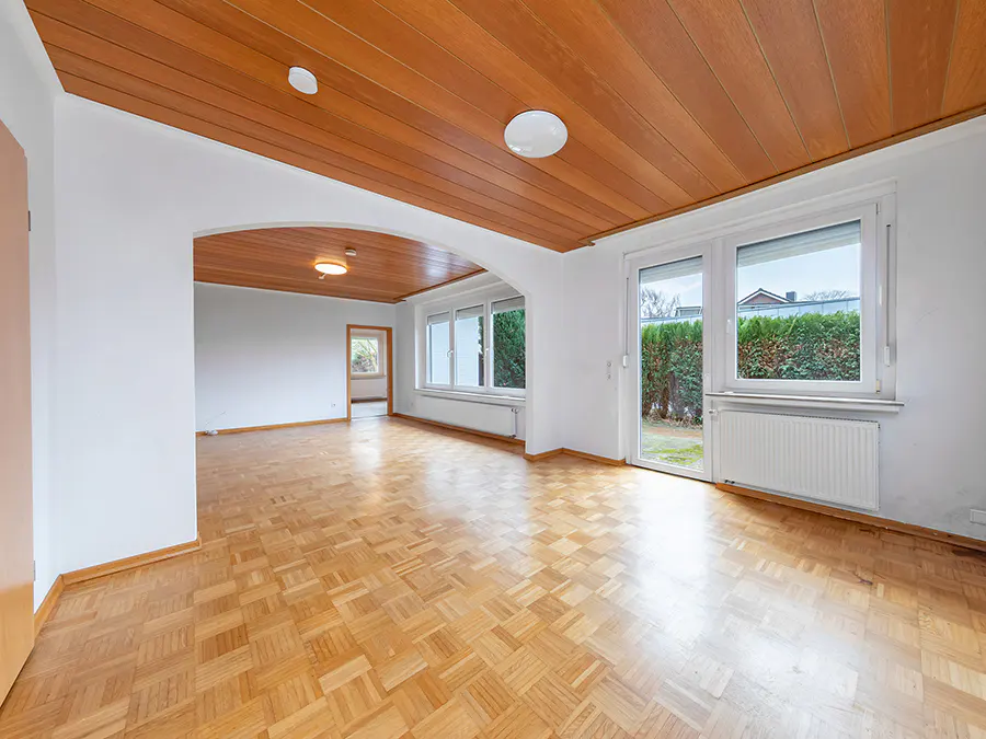 Empty room with parquet floor, white walls, and wood-paneled ceiling. Windows overlook a green hedge.
