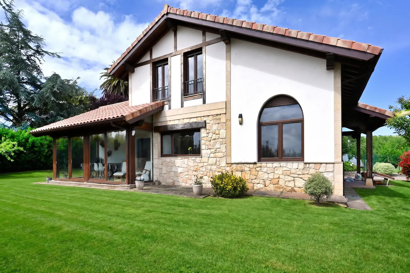 Two-story house with white stucco and stone facade, brown trim, and a red tile roof, surrounded by green lawn.