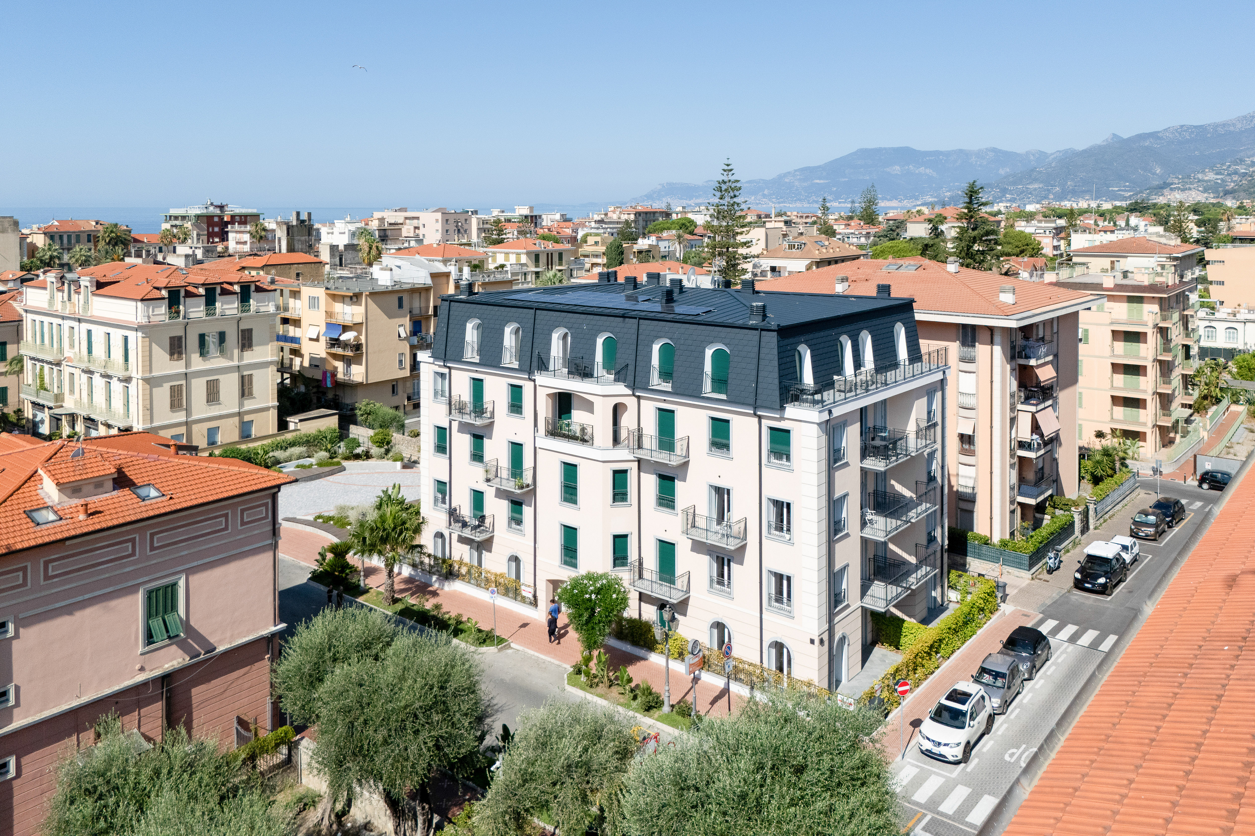 High-angle view of a light-colored apartment building with a dark roof, surrounded by city streets and Mediterranean homes. [AI-generated text]