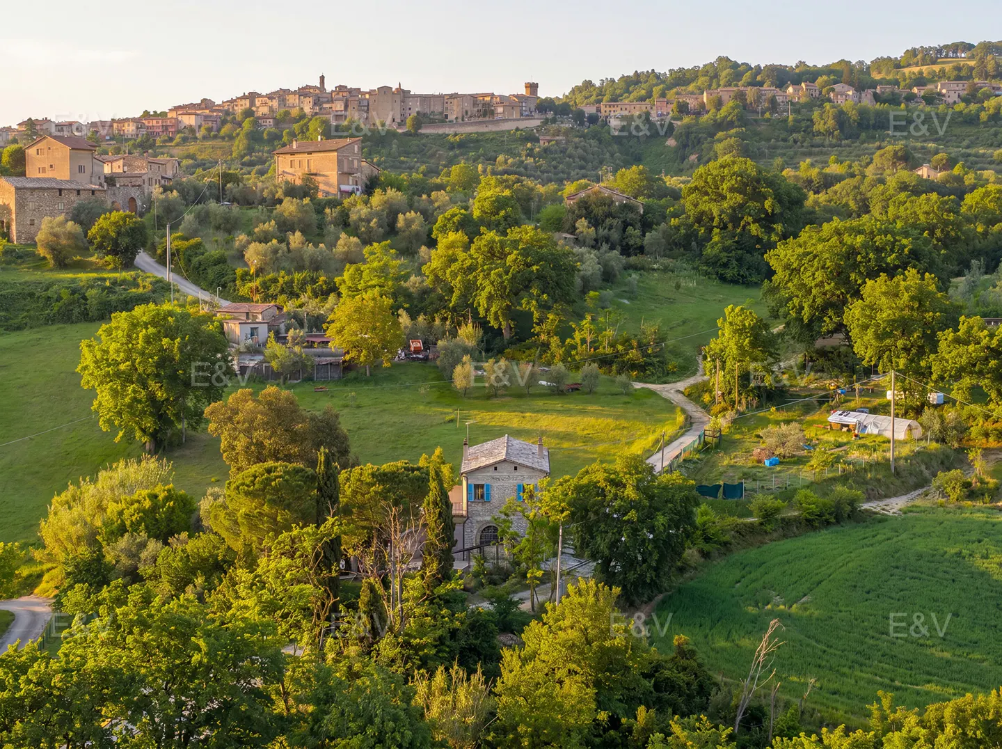 Scenic view of a stone house with blue shutters nestled in green hills, with a village on a distant hilltop.