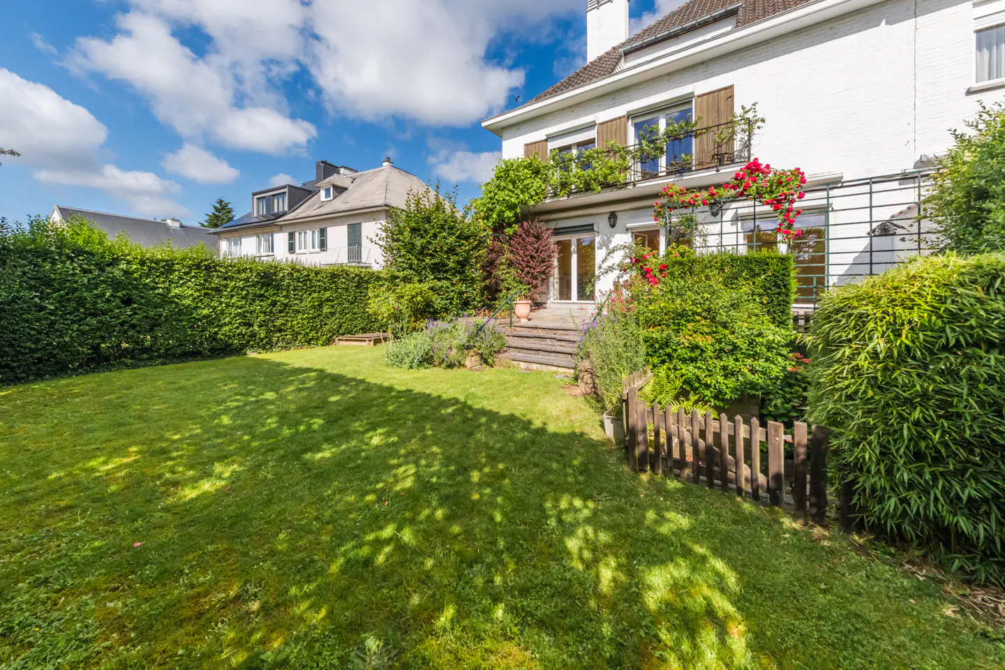 A white house with a lush green lawn, tall hedges, and a small wooden fence under a bright blue sky with white clouds.