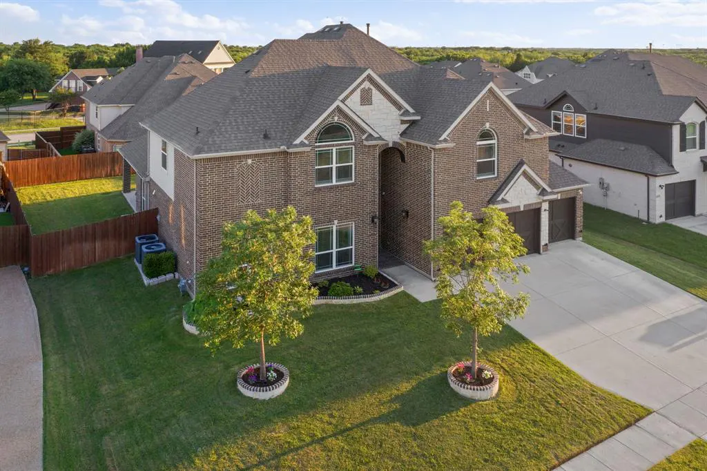 Two-story brick house with a gray roof, green lawn, and two young trees in circular planters.
