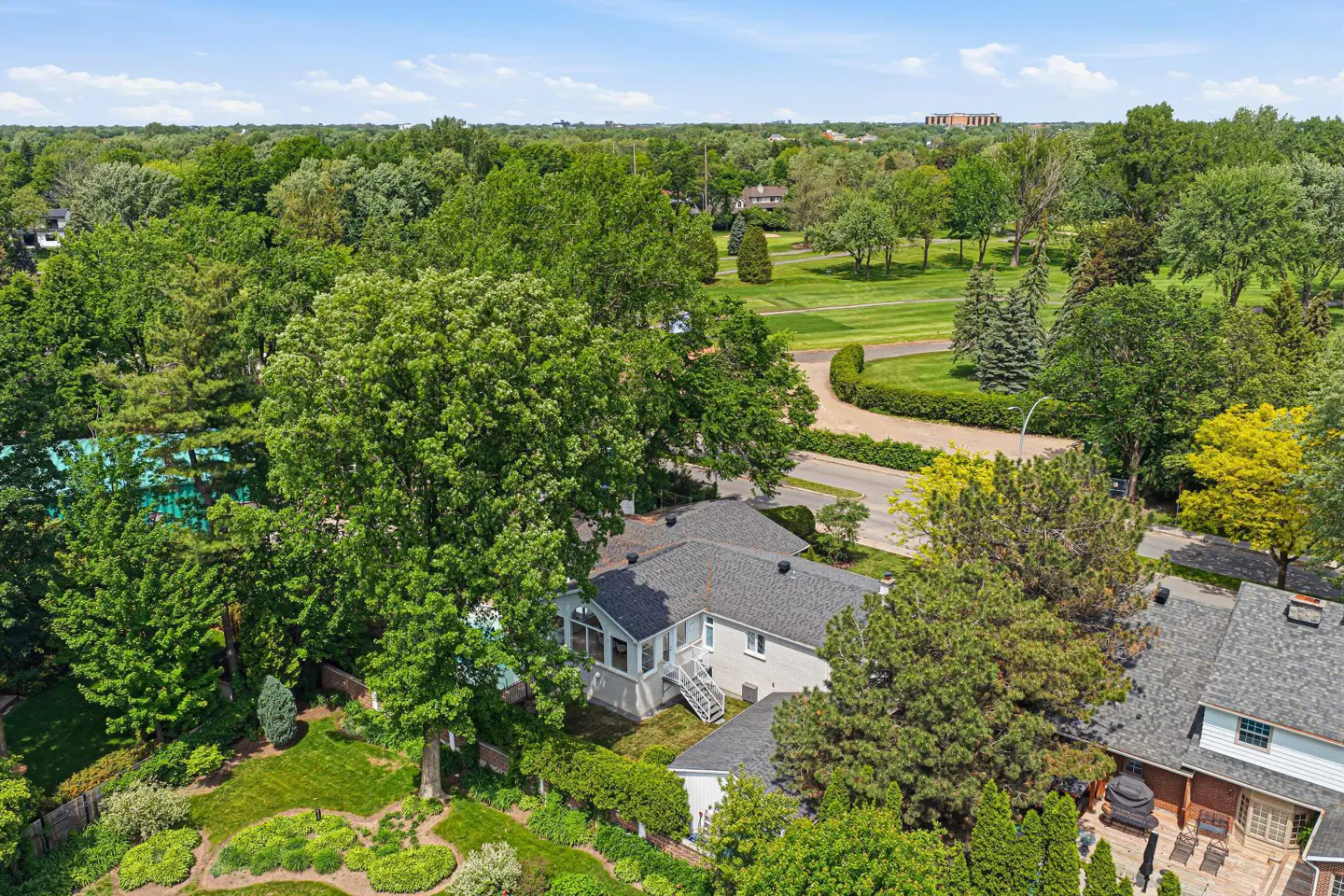 Aerial view of a house with a grey roof nestled among lush green trees and a landscaped garden near a golf course.
