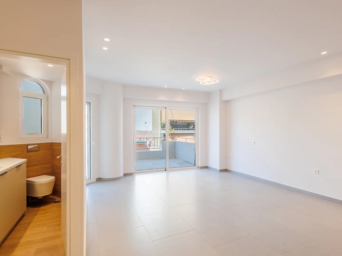 Bright, empty living room with light tile floors, white walls, modern ceiling light, and a view of a balcony.