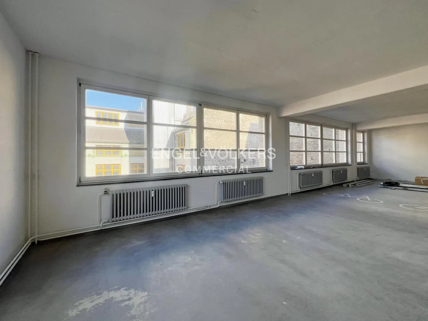 Bright, empty commercial space with large windows, white walls, and gray concrete floors. Radiators line the window wall. Engel & Volkers logo visible.