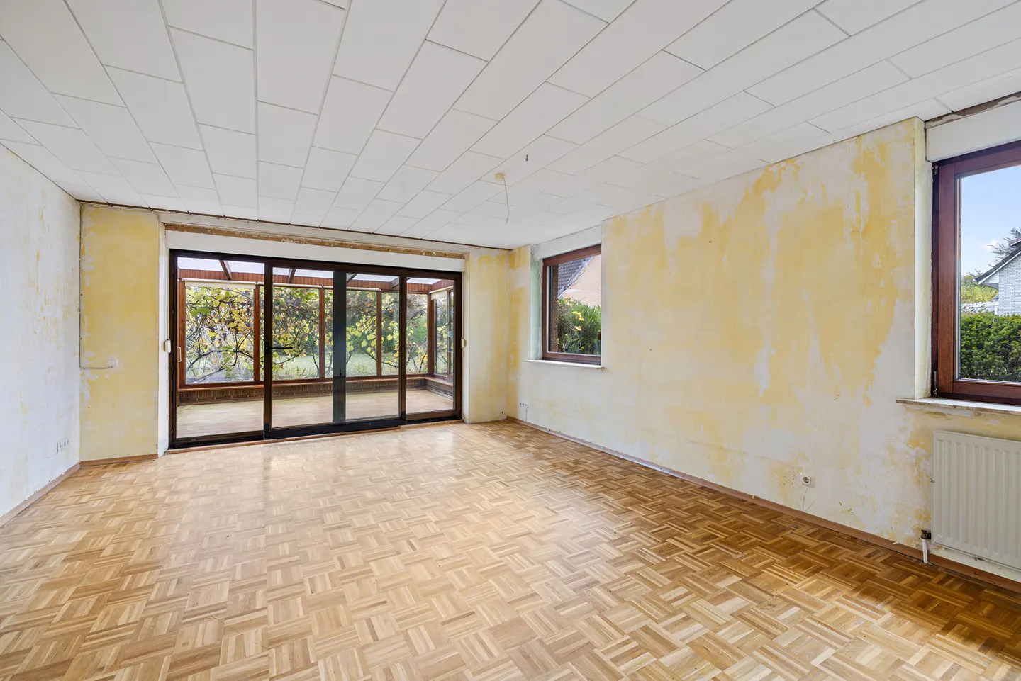 Empty room with parquet floor, peeling yellow paint, and sliding glass doors to a sunroom. Two windows are visible.