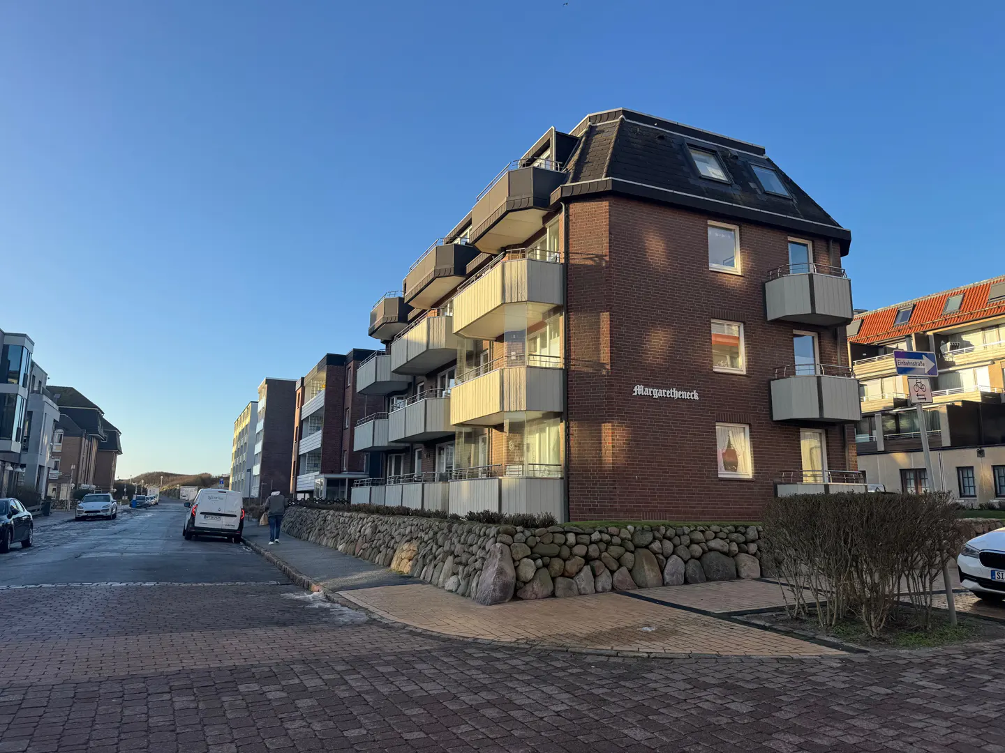 Brick apartment building with white balconies and a dark roof, named "Margaretheneck," sits on a cobblestone street under a blue sky.