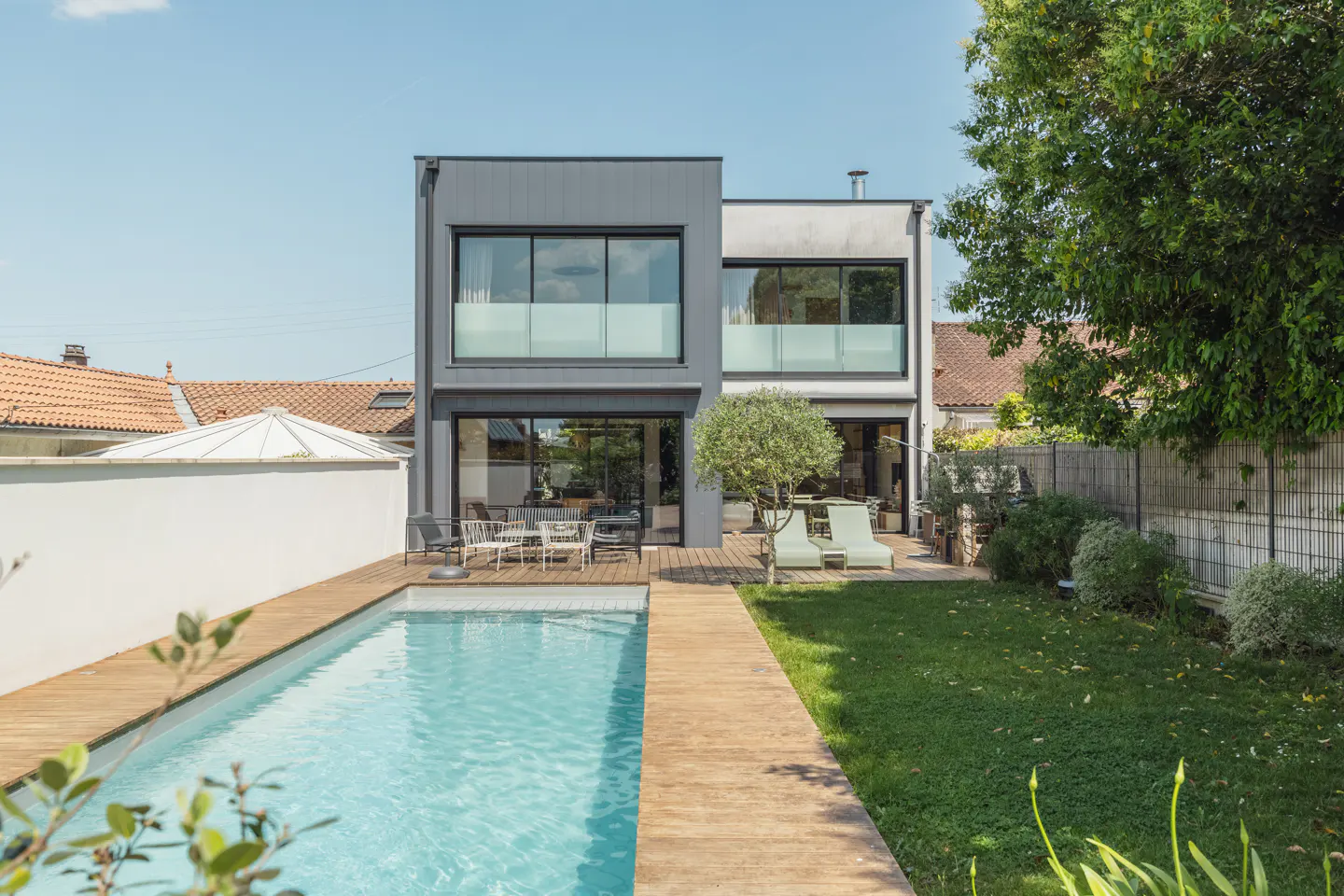 Modern two-story home with a rectangular pool, wooden deck, and green lawn under a clear blue sky.