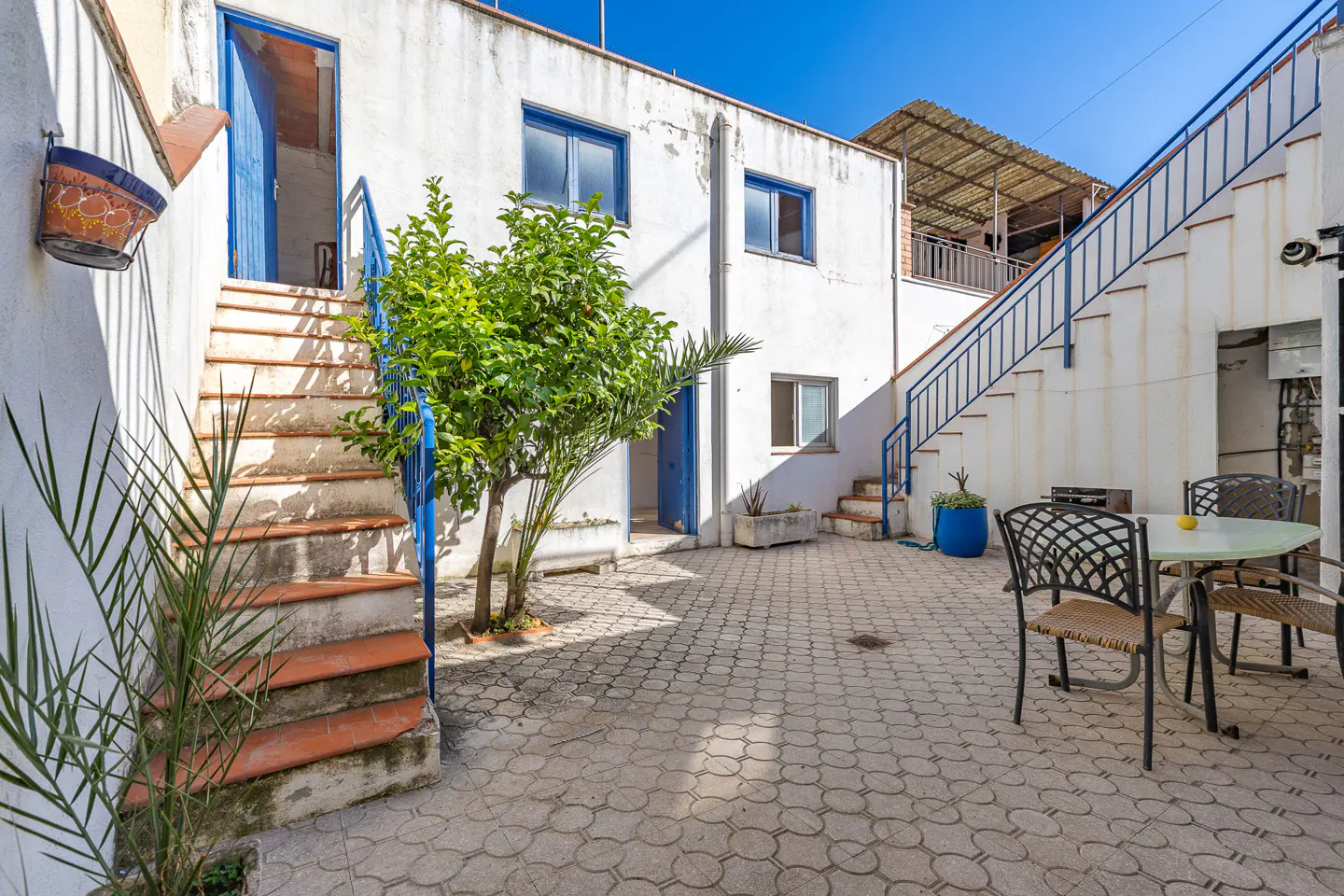 Outdoor courtyard with white buildings, blue trim, and patterned stone floor. Stairs lead to upper levels. Table and chairs are on the right.