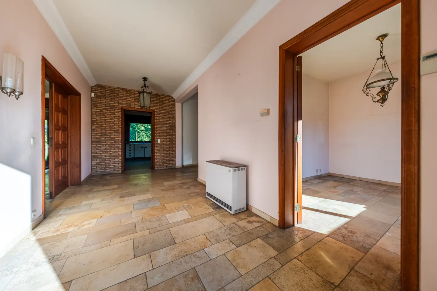 Hallway with light pink walls, stone tile flooring, a brick accent wall, and doorways leading to adjacent rooms.