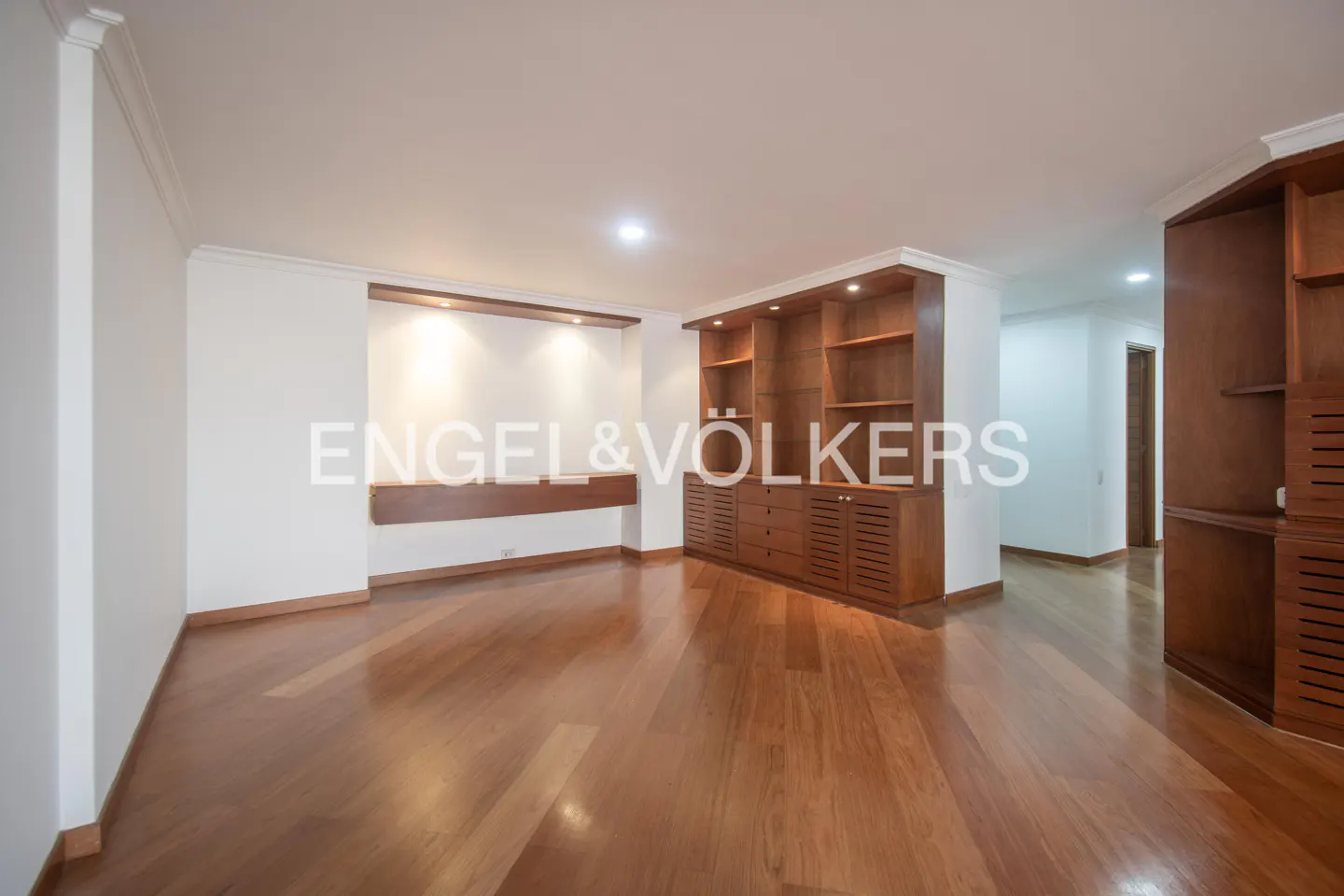 Empty living room with hardwood floors, white walls, and built-in wooden shelving and cabinets.