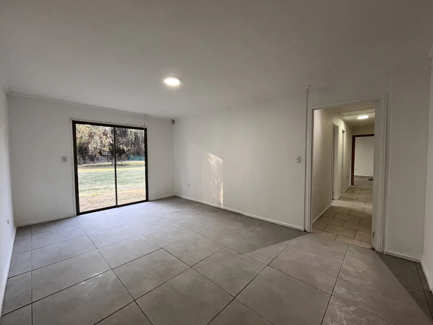 Bright, empty room with white walls, gray tile floor, and sliding glass doors to a green yard. Hallway visible to the right.