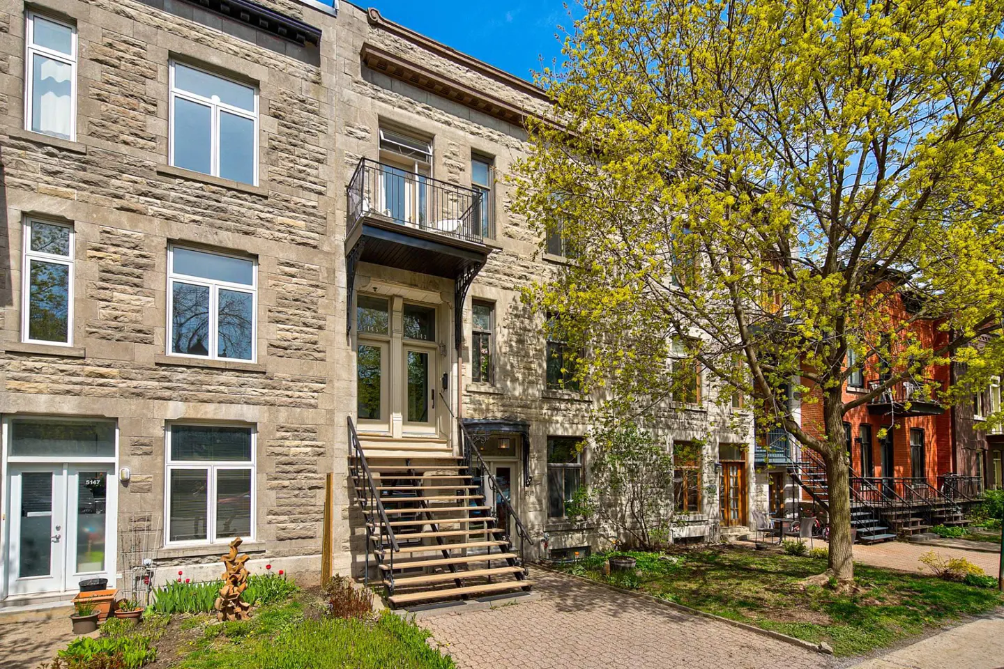Row of stone townhouses with black iron stairs and balconies, and a large tree with yellow-green leaves.