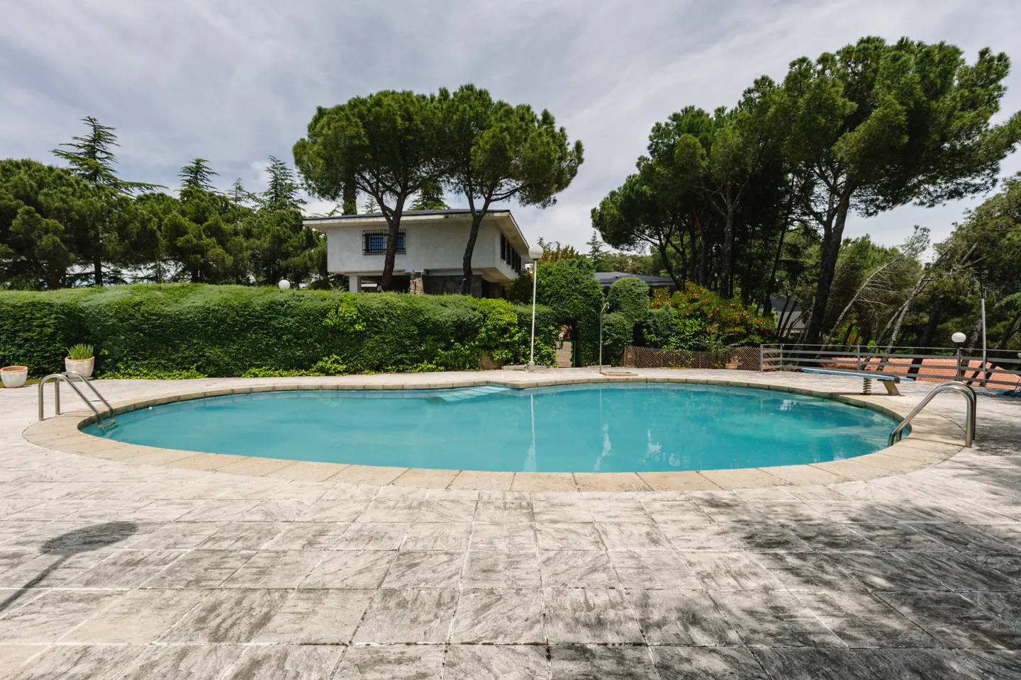 Outdoor pool with turquoise water, surrounded by a stone patio. A white house is visible behind green hedges and trees.