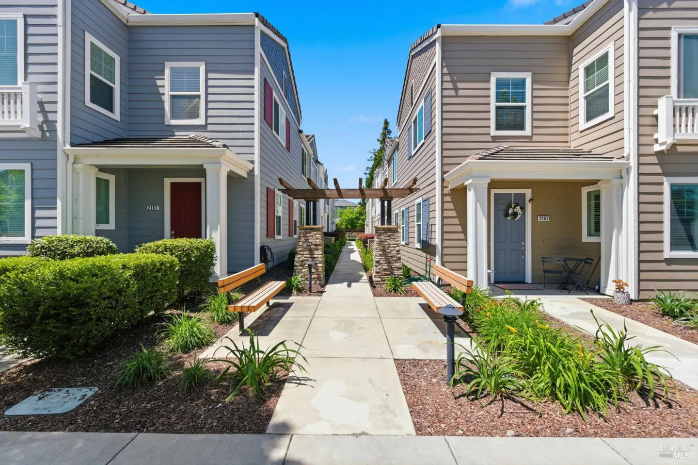 Exterior view of two-story townhouses with a walkway, benches, and pergola on a sunny day.