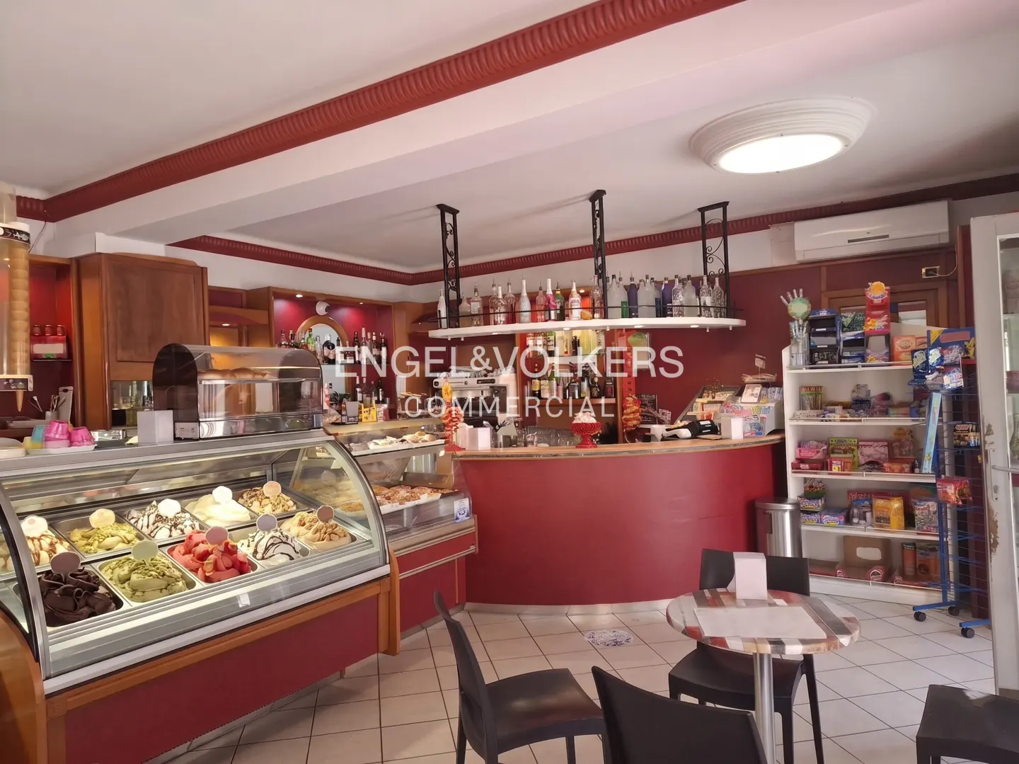 Interior view of a commercial space with gelato display case, red counter, tables, and shelves stocked with snacks.