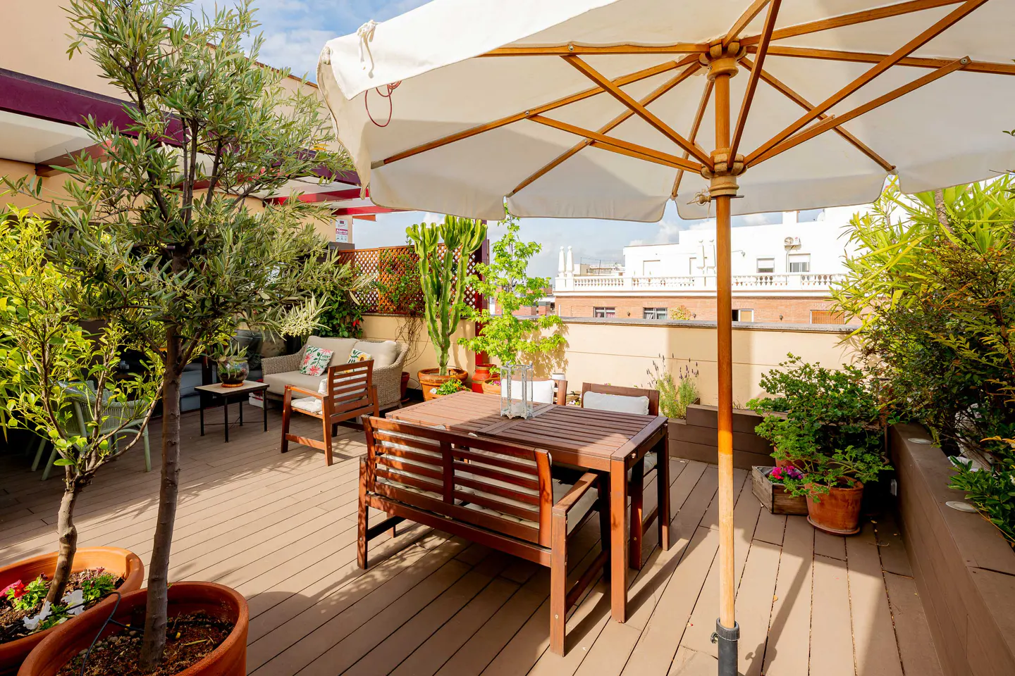 A rooftop patio with a wooden table and chairs under a white umbrella, surrounded by potted plants and a view of city buildings.