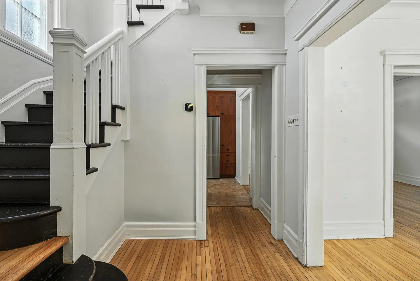 Hallway with hardwood floors, white walls, and black and white staircase. Kitchen visible through doorway.