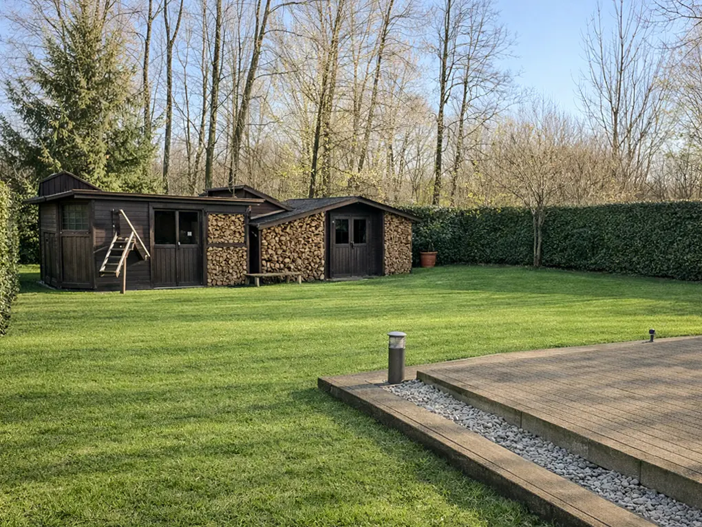 A backyard with a green lawn, a dark brown shed with a staircase, and a wood storage area.