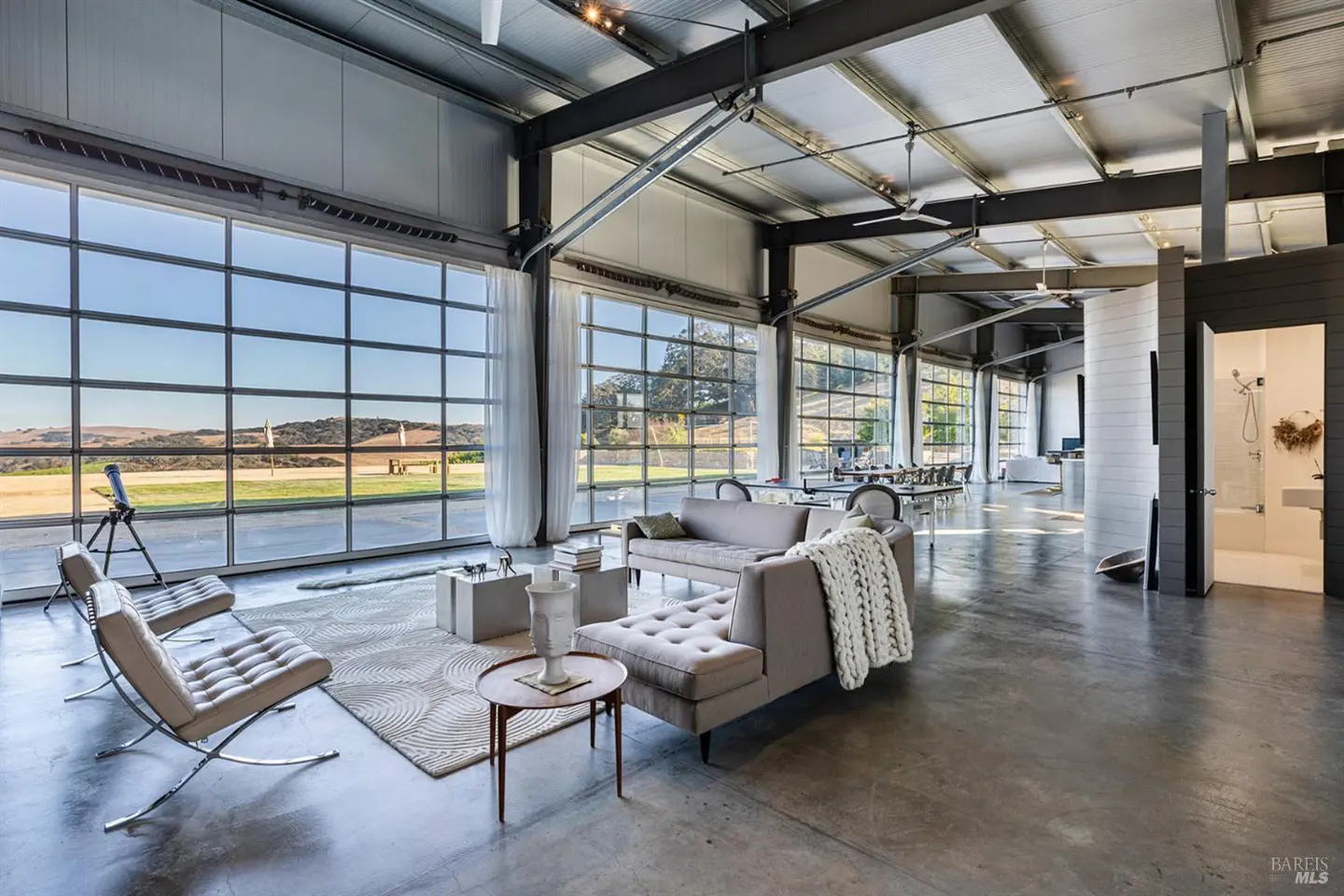 A modern living room with large windows, gray sofas, and a view of rolling hills.