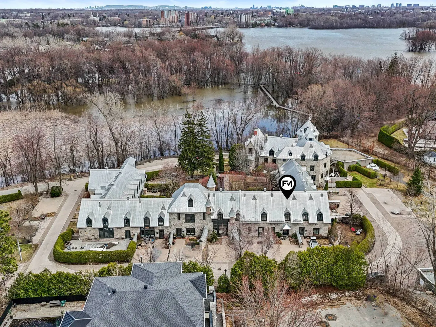Aerial view of grey stone townhouses with white roofs, surrounded by trees and water. City skyline in the distance.