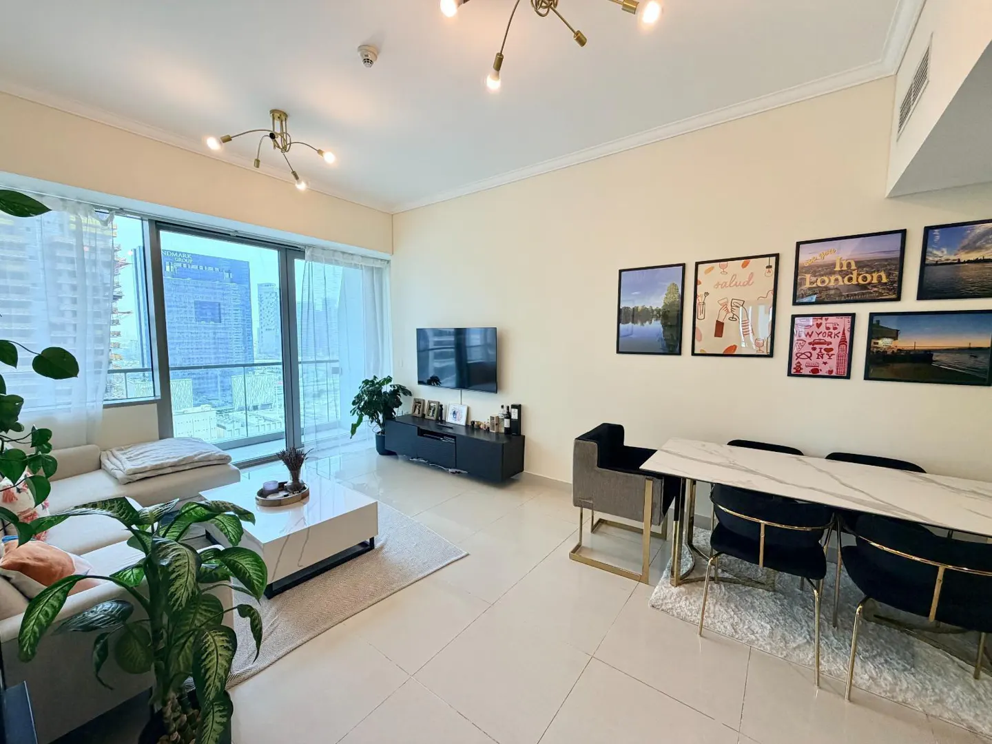 Bright living room with a white sofa, marble table, and city view through a large window. Artwork adorns the wall above a dining area.