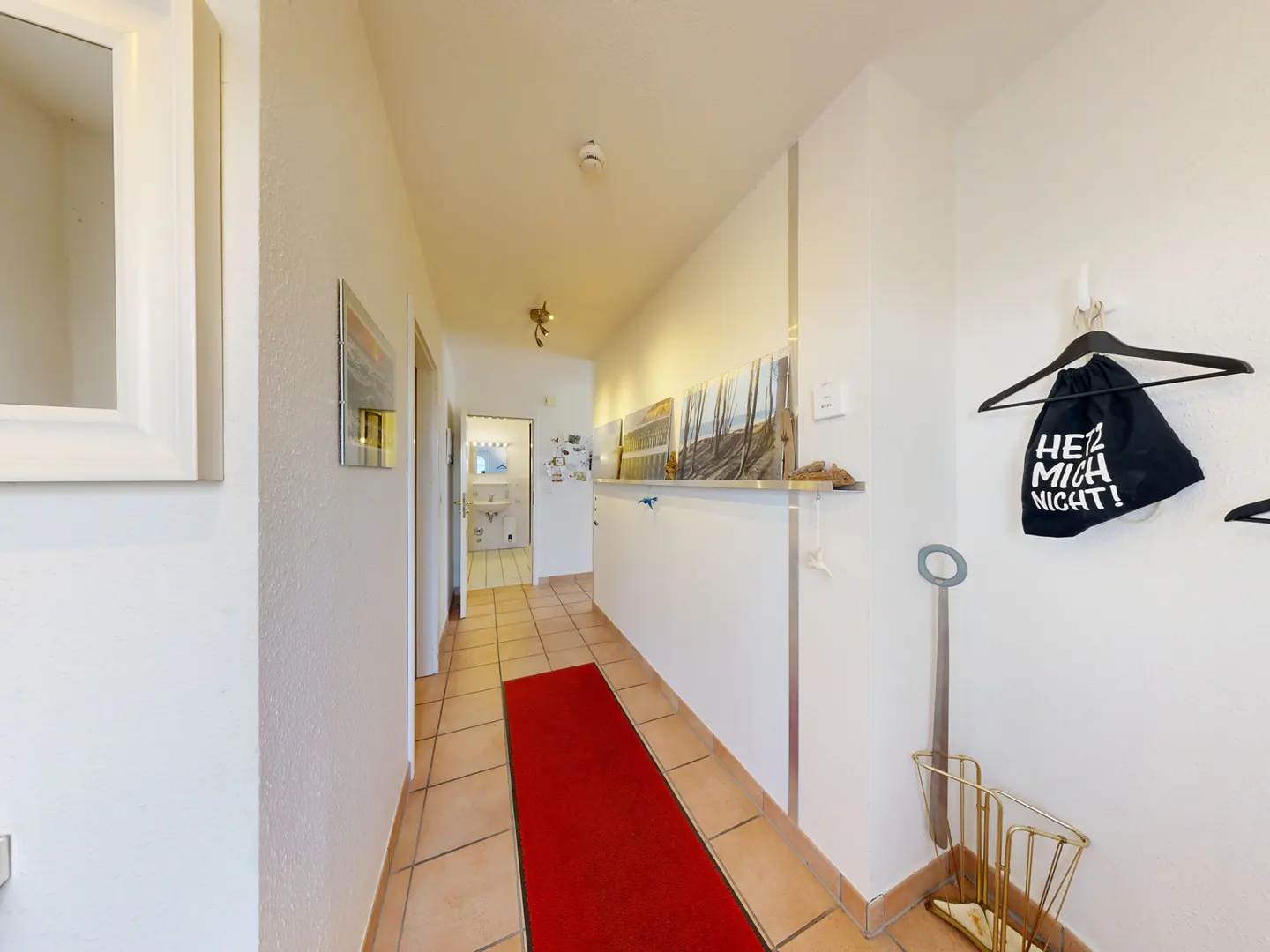 Hallway view with a red runner rug on tiled floor. White walls with art, coat rack, and umbrella stand. Bathroom visible in the background.