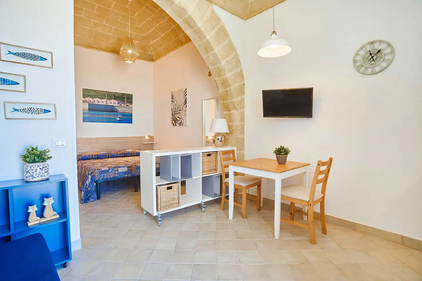 Bright studio apartment with stone archway, bed, dining table, TV, and blue shelving unit.