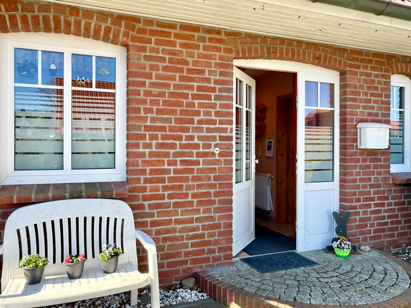 Exterior of a red brick house with an open white door, a white bench with flowers, and a stone walkway.