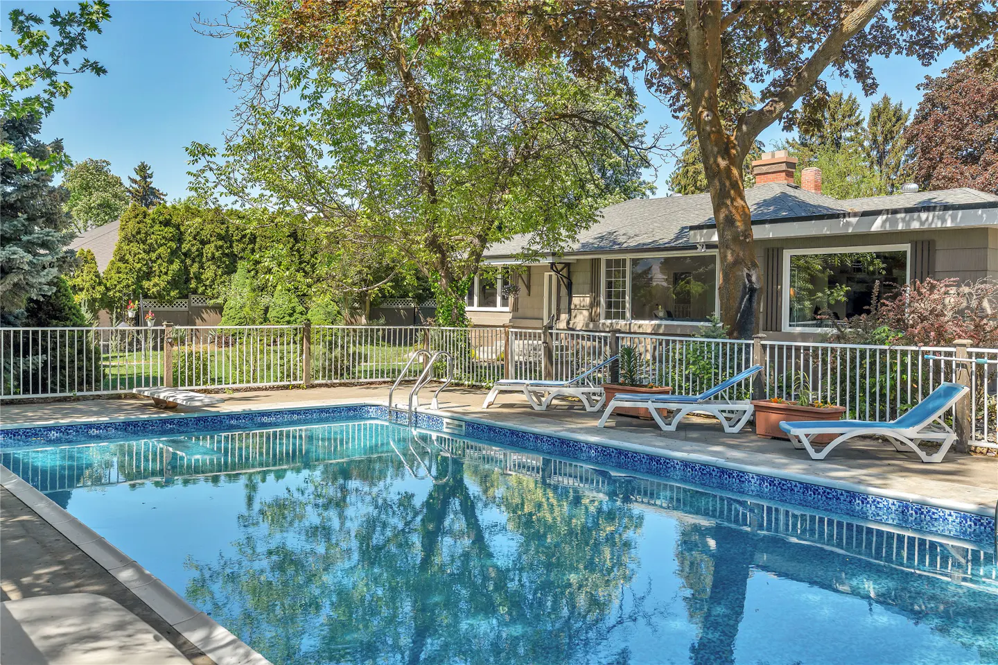 A backyard pool with blue water reflecting trees, surrounded by a white fence, lounge chairs, and a house.