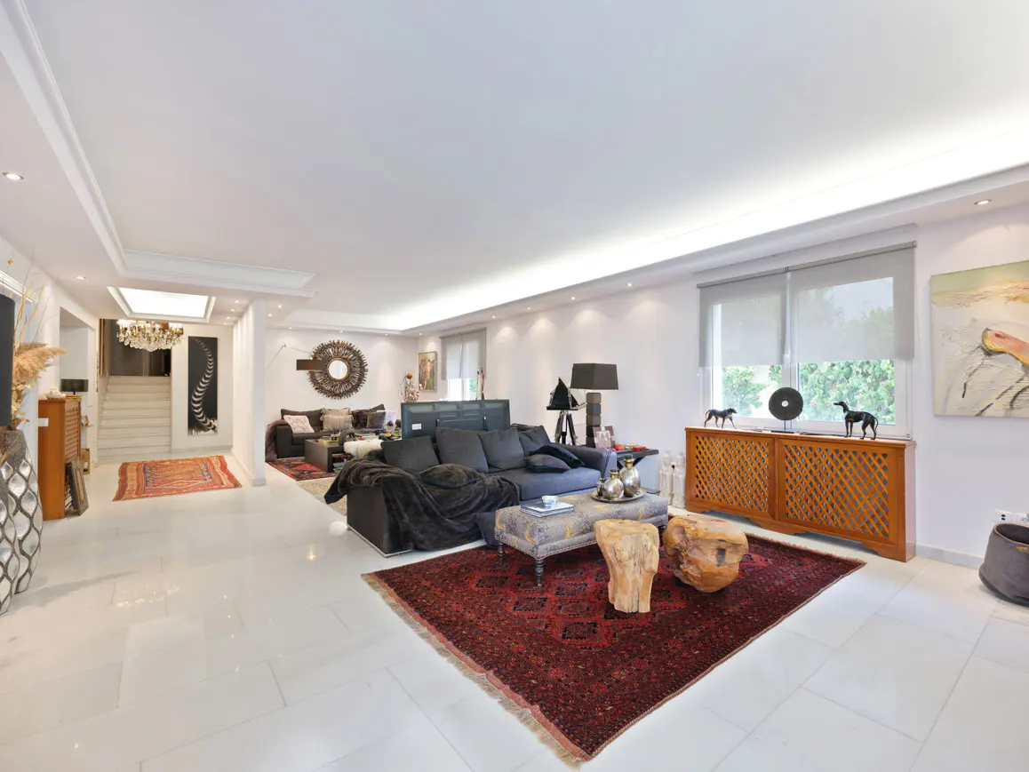 Spacious living room with white floors, a large dark sofa, a red patterned rug, and wooden accent tables.