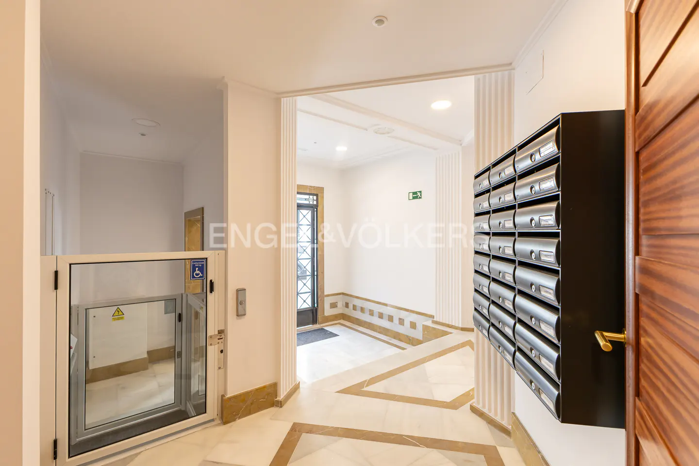 Hallway with marble floors, mailboxes, and a wheelchair lift. The walls are white, and the mailboxes are silver. A wooden door is on the right.