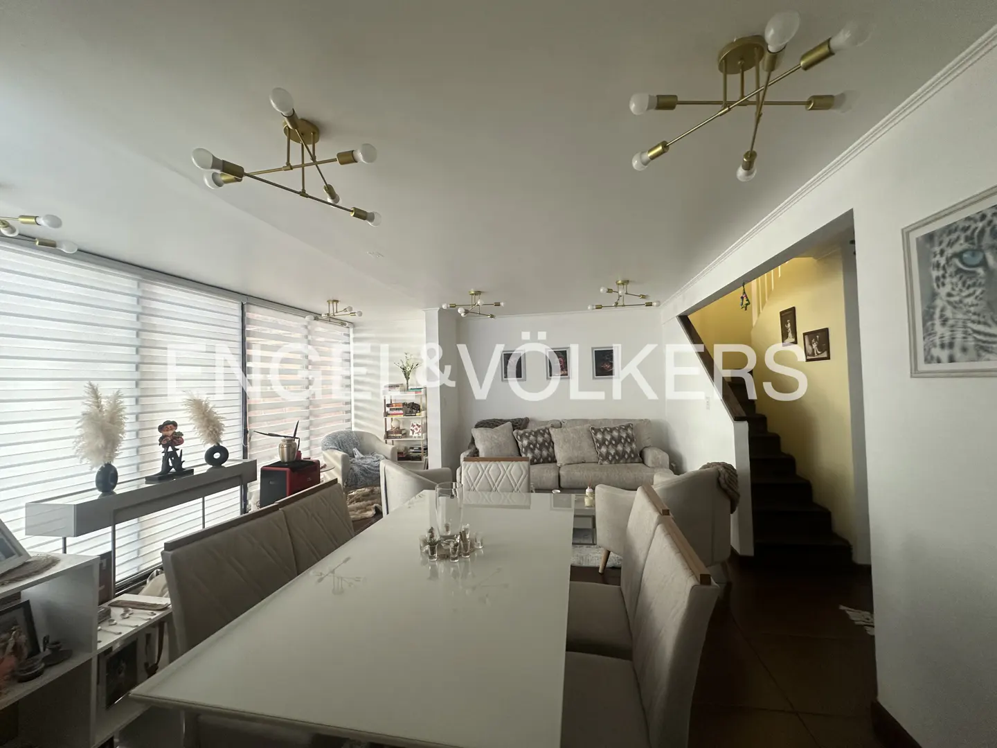Bright, modern living and dining area with white table, beige chairs, and a gray sofa. Gold light fixtures adorn the ceiling. Stairs lead to the upper level.
