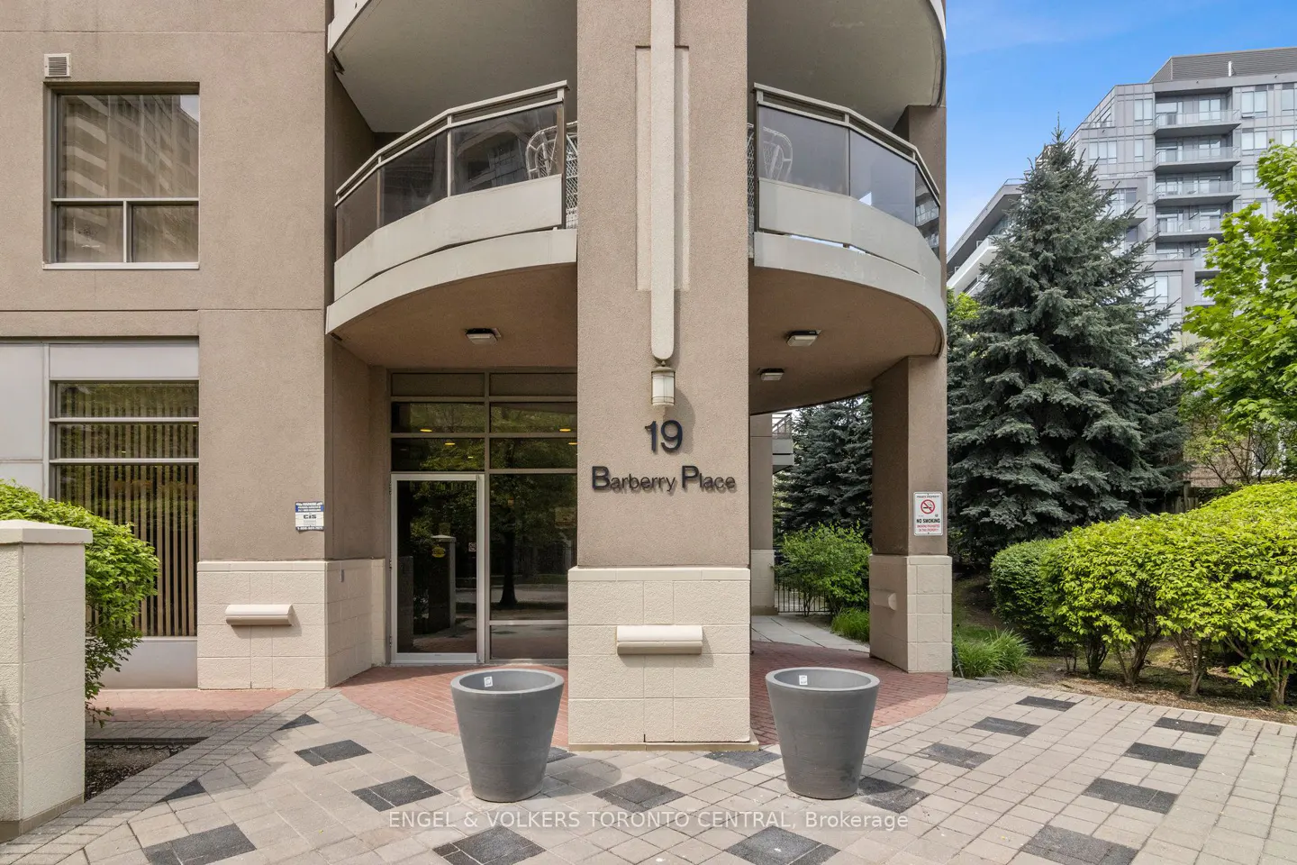 Exterior of 19 Barberry Place, a beige condo building with a curved balcony. Two gray planters flank the entrance on a patterned brick walkway.
