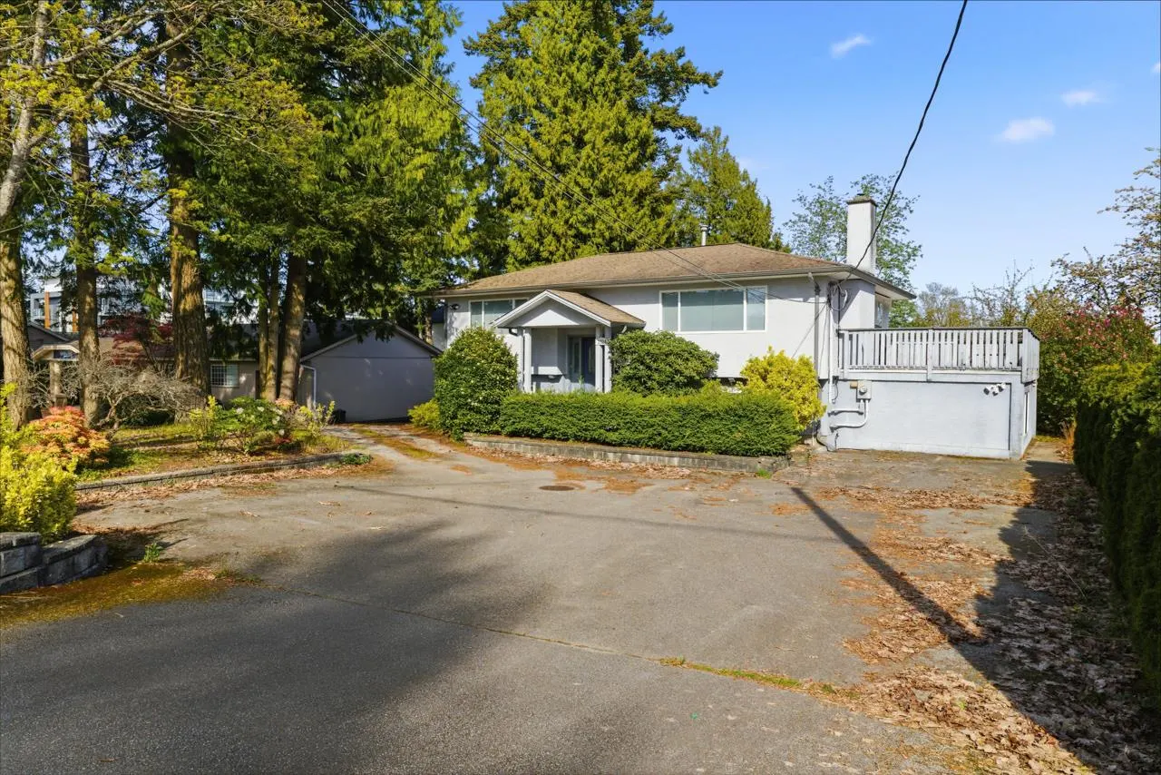 A single-story light gray house with a deck, surrounded by green trees and bushes.