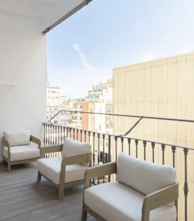 Balcony with three white cushioned chairs, a black railing, and a city view under a blue sky.