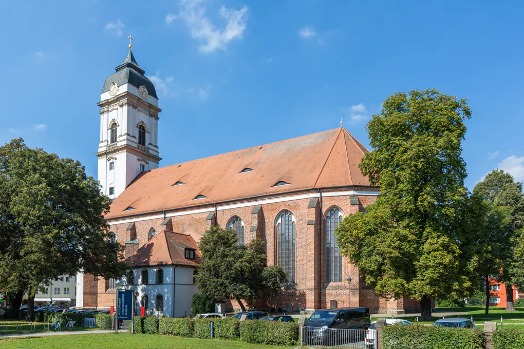 A historic red-brick church with a tall white tower and orange tiled roof stands under a clear blue sky, framed by trees.