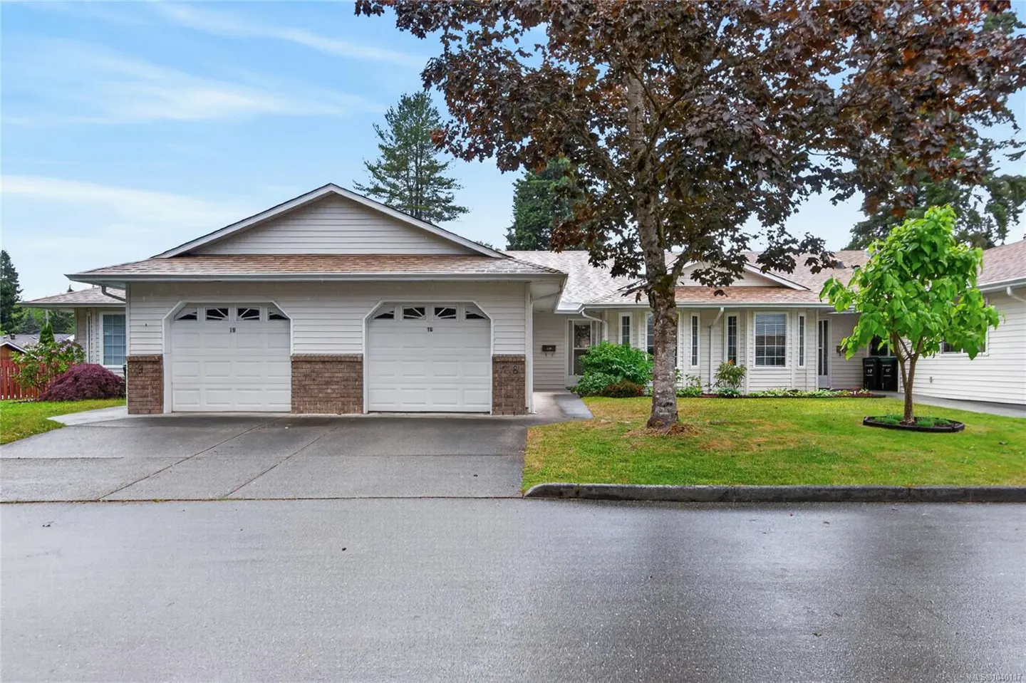 Single-story duplex with white siding, two white garage doors, a brown roof, and a front lawn with trees under a blue sky.