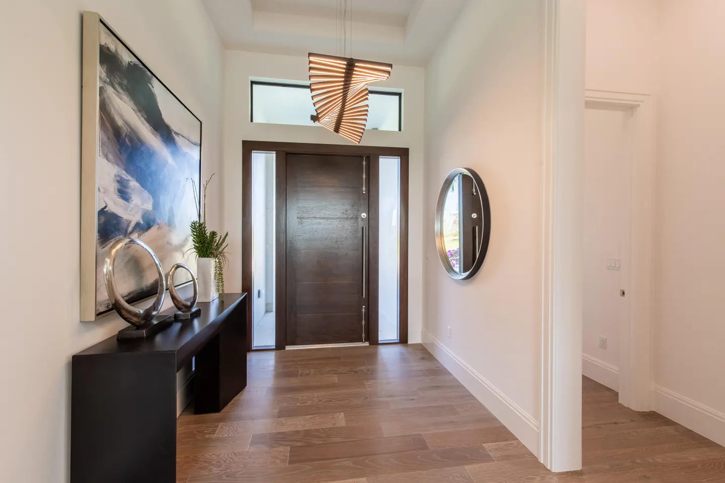Foyer with a dark wood door, light wood floors, and white walls. A black console table with art and decor is on the left. A round mirror hangs on the right.