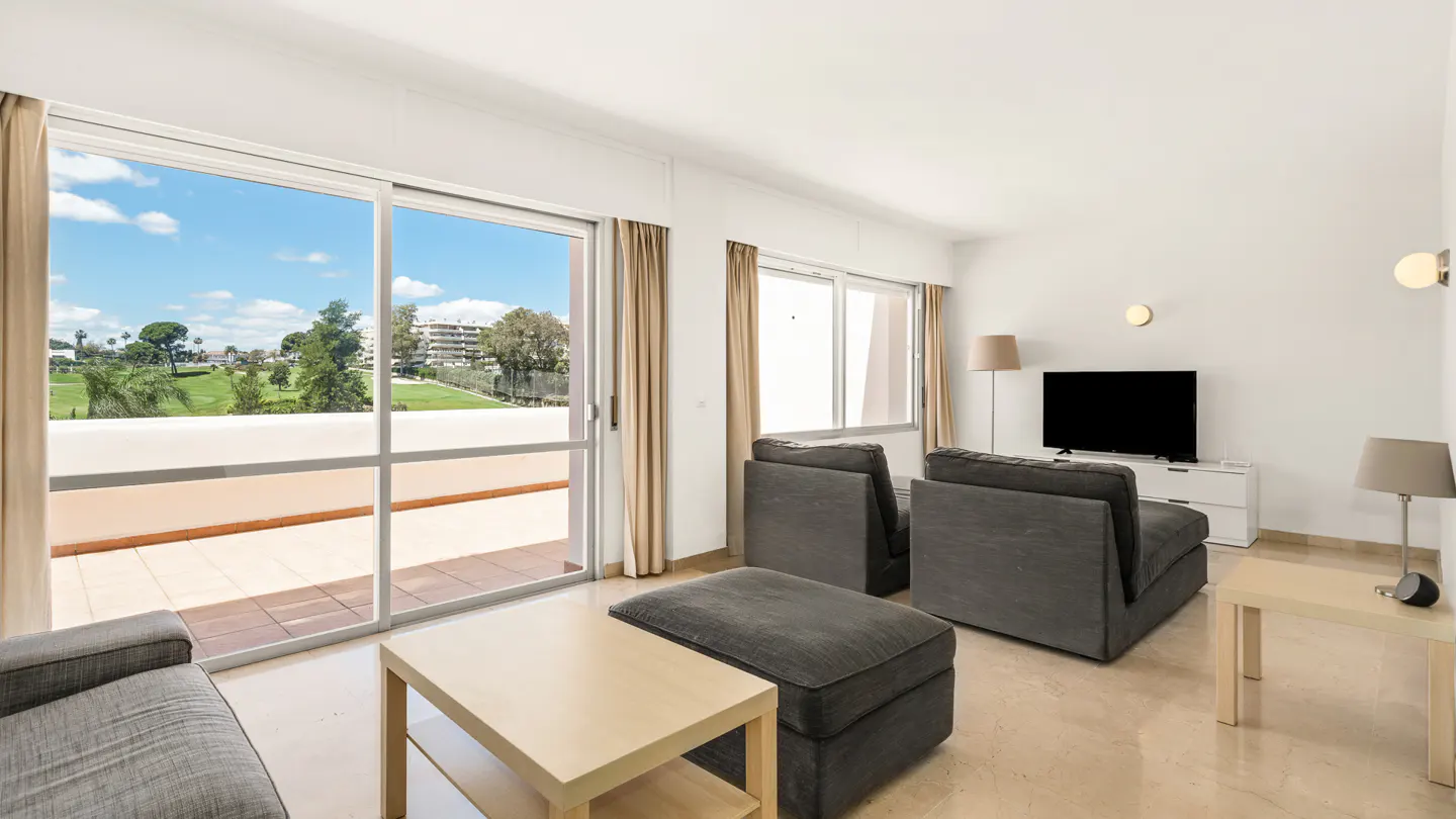 Bright living room with gray sofas, light wood tables, and a TV. Sliding glass doors open to a balcony with a view of a green landscape.
