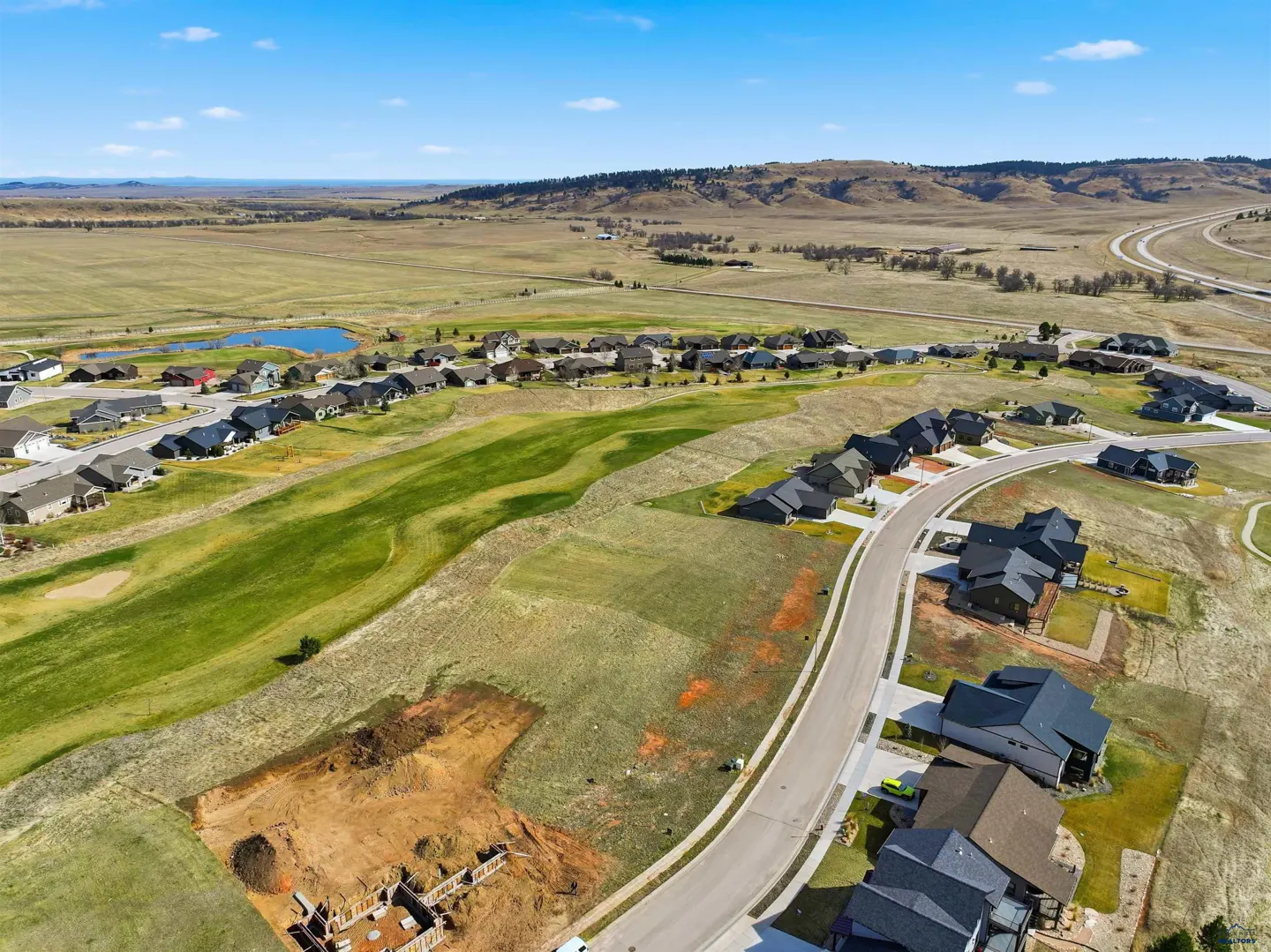 Aerial view of a residential area with houses, green spaces, and a road winding through the neighborhood. In the background, there's a vast landscape with hills and a blue sky.