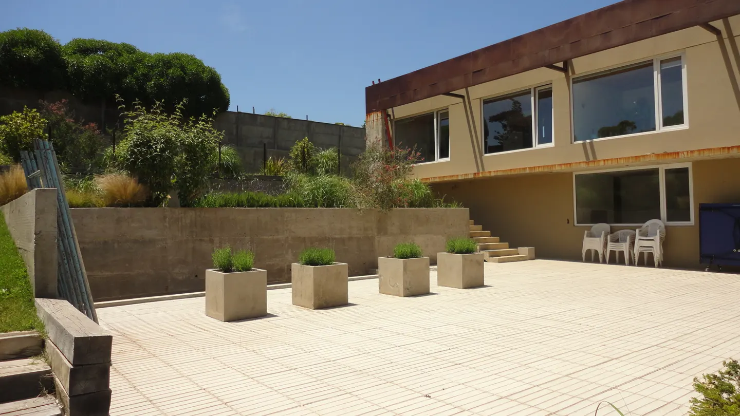 Exterior view of a beige house with a tiled patio, four square planters, and a retaining wall with greenery. Blue sky.
