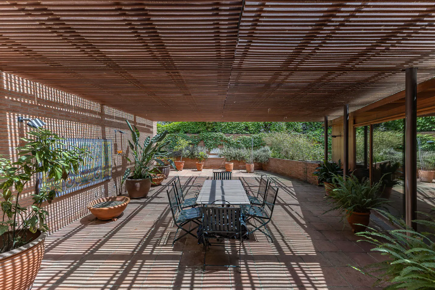 Outdoor patio with a long table and chairs under a slatted wooden roof, casting shadows on the brick floor. Green plants and foliage surround the space.