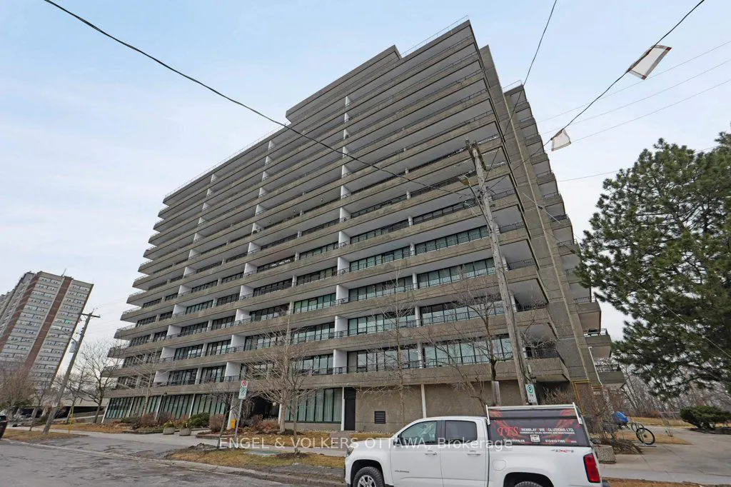 Exterior view of a tall, gray apartment building with many balconies, a white truck parked in front, and trees nearby.