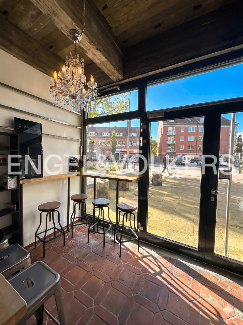 Interior view of a cafe with stools, a counter, and a chandelier. Large windows overlook a sunny outdoor plaza.