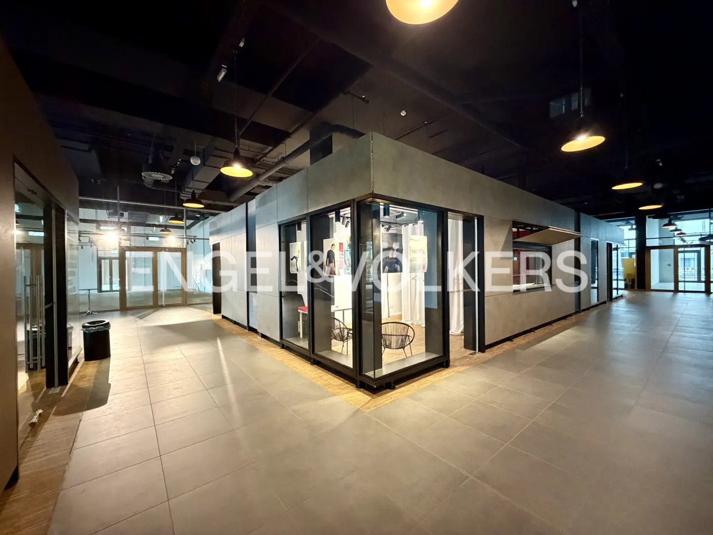 Engel & Volkers real estate office with glass walls, gray tile floor, and black ceiling with hanging lights.