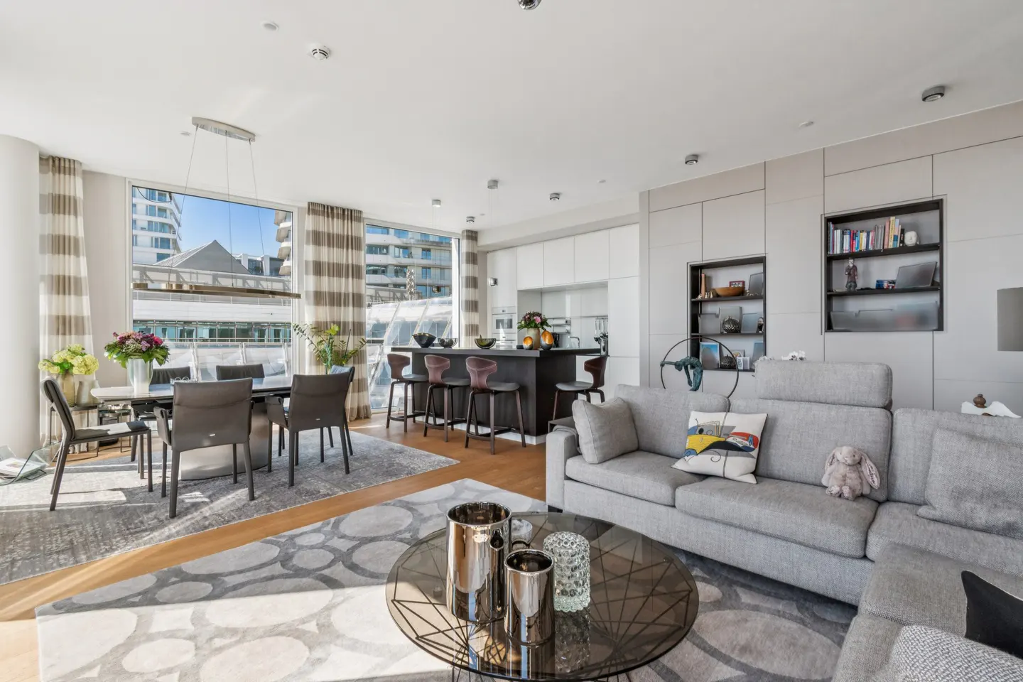 Bright, modern open-concept living space with a gray sofa, dining table, kitchen island, and large windows.